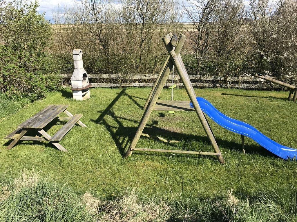 Un parc infantil amb un gronxador de fusta, un tobogan blau i una taula de picnic en una zona verda amb arbres al fons.