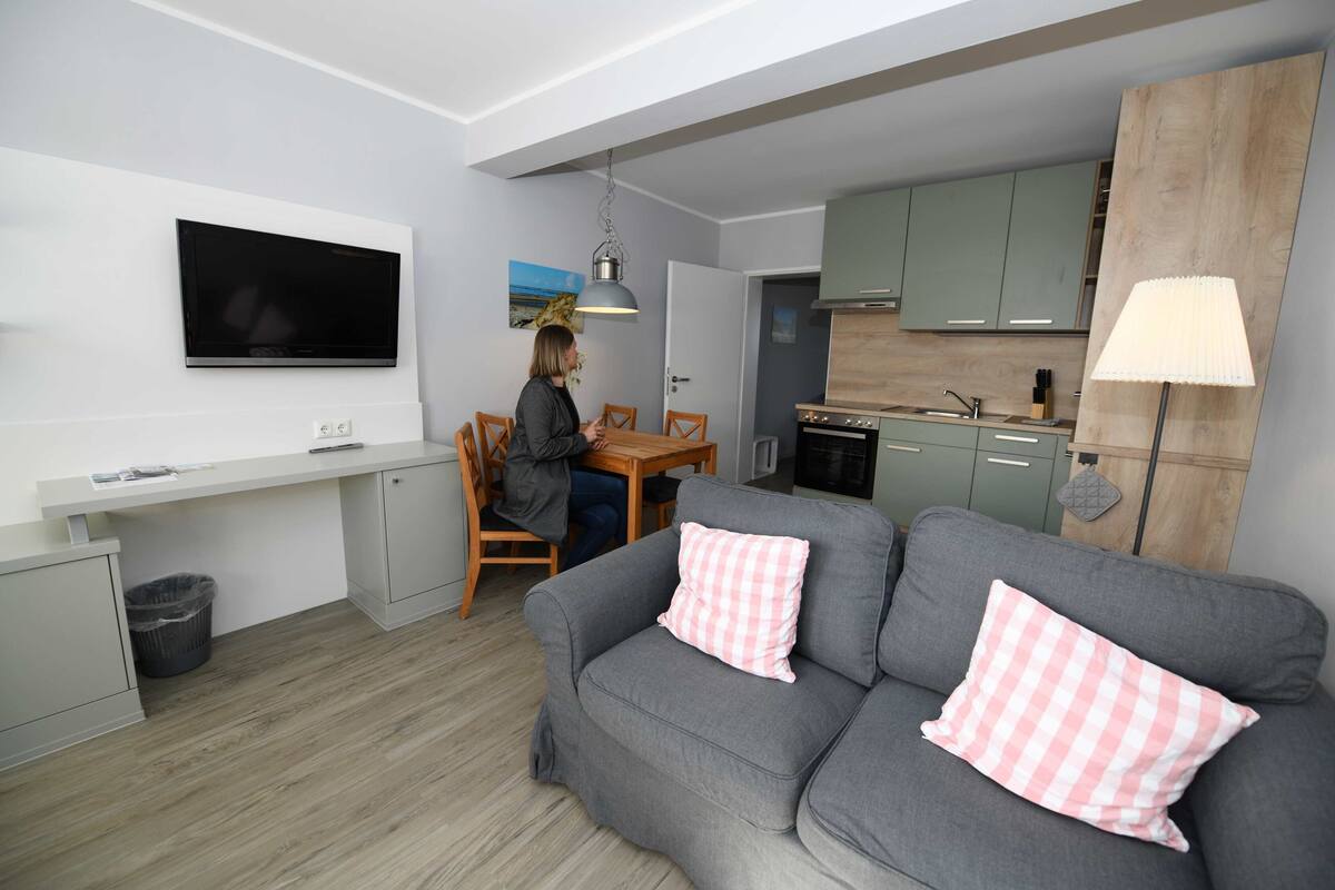 A modern living space with a grey sofa, a dining table, and a kitchen area.