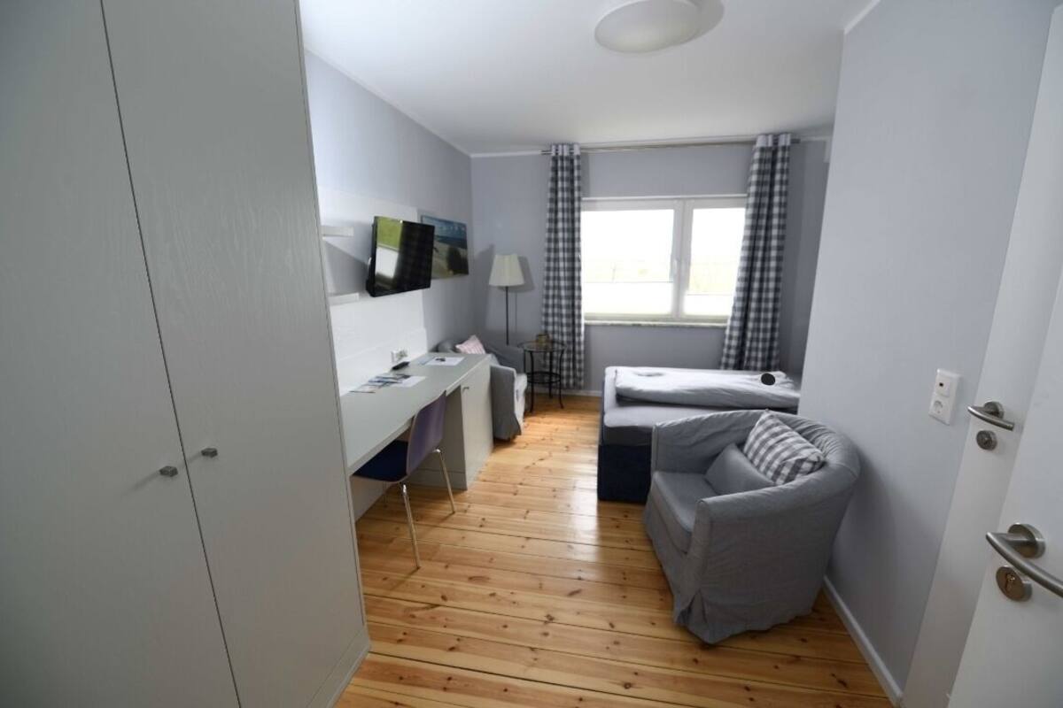 A compact room with a wooden floor, a desk with a chair, a bed, and a window with curtains.