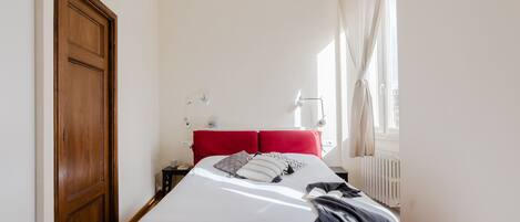 Un dormitorio con puerta de madera, cama con ropa de cama roja, mesita de noche, lámpara y ventana con cortinas.
