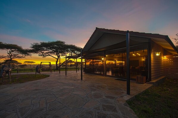 A modern outdoor seating area with stone flooring, a covered patio, and a view of the sunset. - Serengeti Sametu Camp (Serengeti)