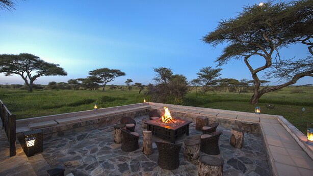 An outdoor seating area with a fire pit, stone flooring, and wooden stools. - Serengeti Sametu Camp (Serengeti)
