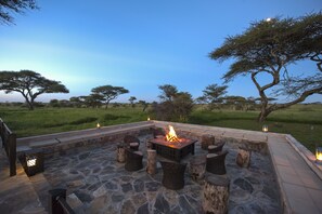An outdoor seating area with a fire pit, stone flooring, and wooden stools. - Serengeti Sametu Camp (Serengeti)