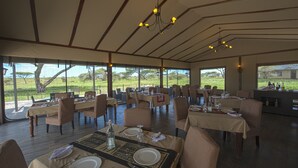 An indoor dining area with tables set for meals, large windows offering views of the outdoors, and a chandelier hanging from the ceiling. - Serengeti Sametu Camp (Serengeti)