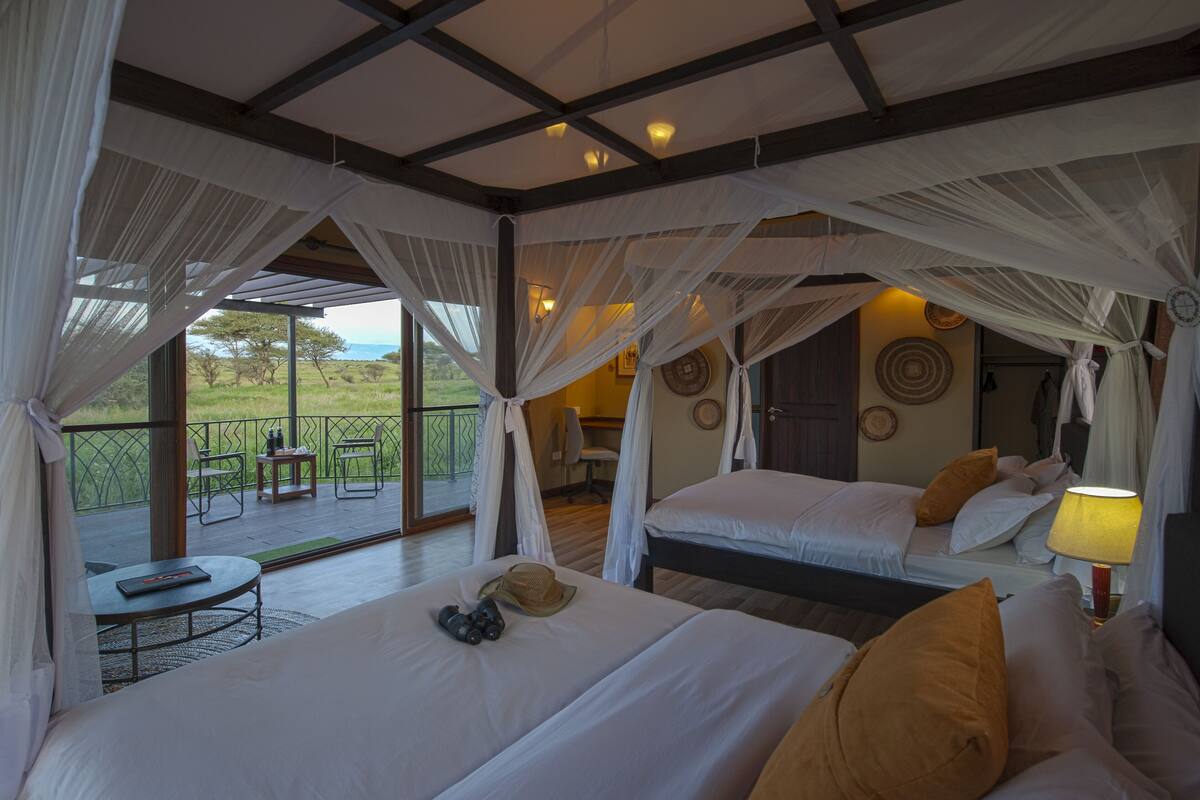 A room with two beds, a table, and a chair, with a view of the outdoors.