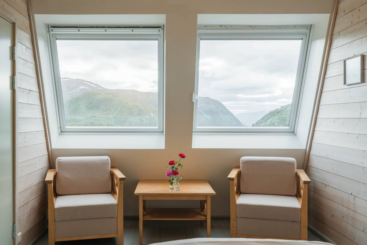 A room with two windows offering a mountain view, a small table with a vase of flowers, and two chairs.