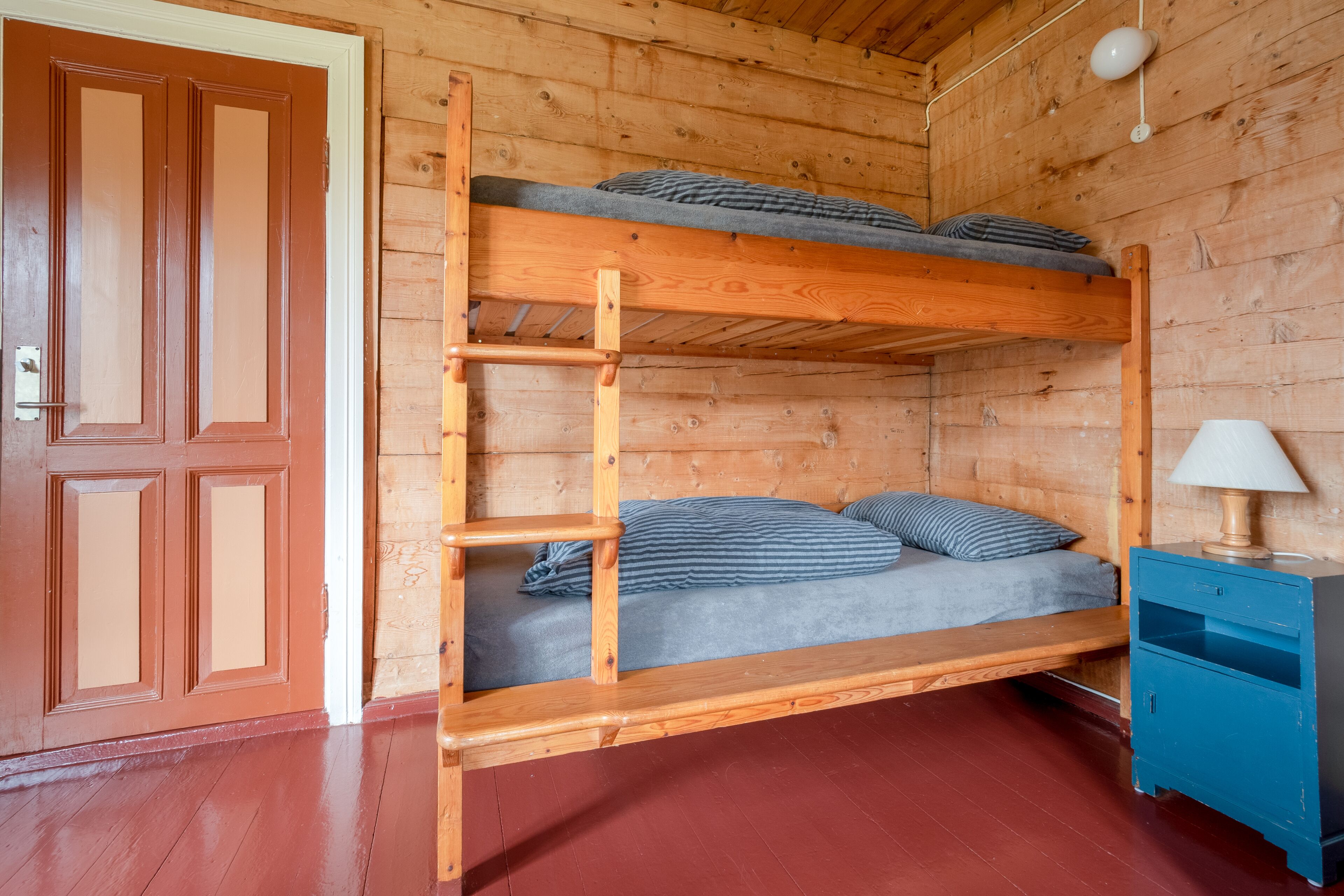 A wooden bunk bed with a ladder, a bedside table with a lamp, and a door with a handle.