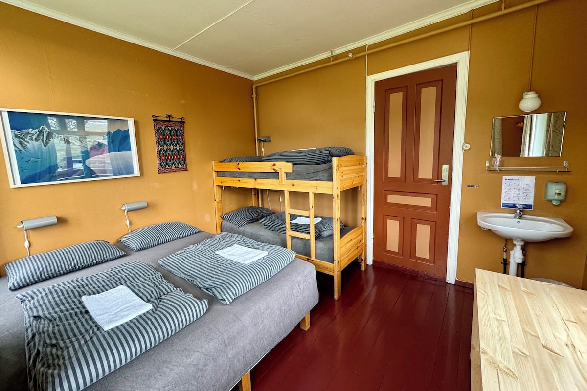 A dormitory room with bunk beds, a single bed, a wooden desk, a sink, and a mirror.