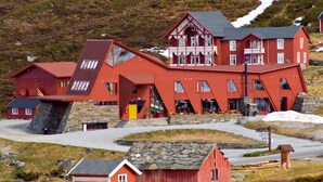 A red wooden building with multiple windows and a stone base, located in a rural area with a road and a small wooden structure nearby. - Turtagrø Hotel (Luster)