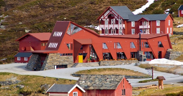 A red wooden building with multiple windows and a stone base, located in a rural area with a road and a small wooden structure nearby. - Turtagrø Hotel (Luster)