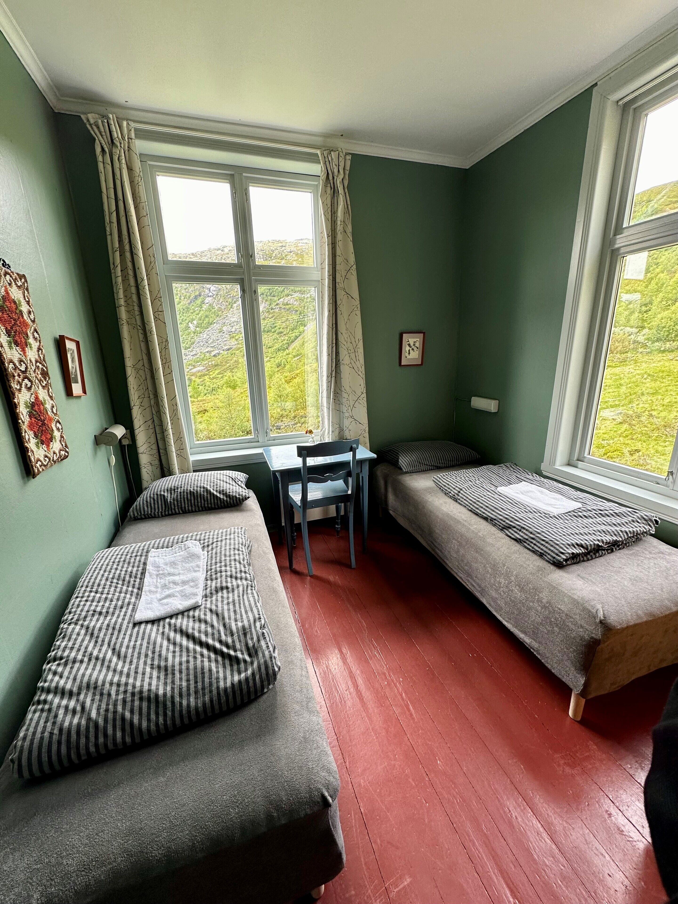 A room with two beds, a desk, and a window with a view of green hills.