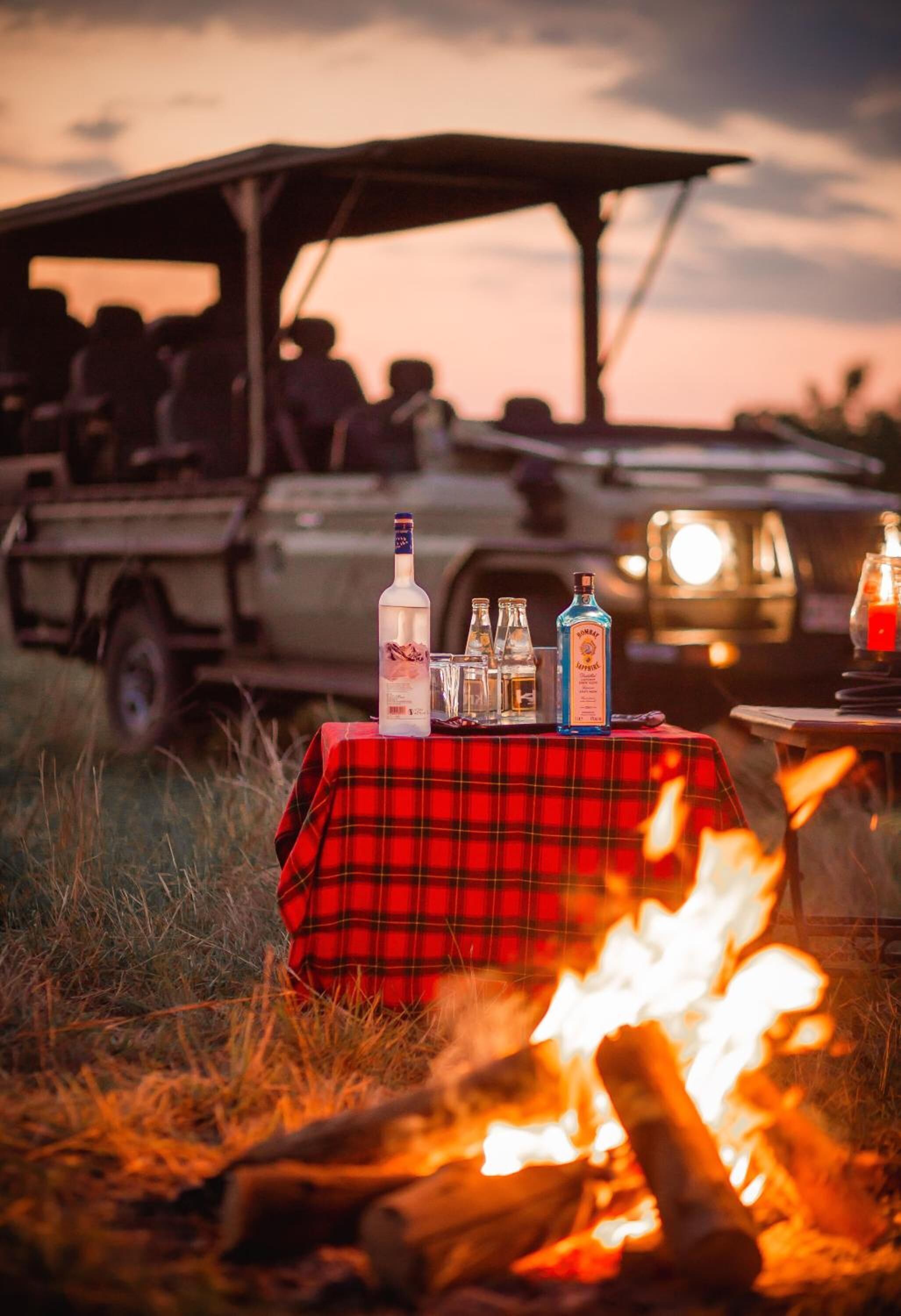 A table with bottles and glasses set up outdoors with a fire in the foreground.