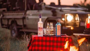 A table with bottles and glasses set up outdoors with a fire in the foreground.