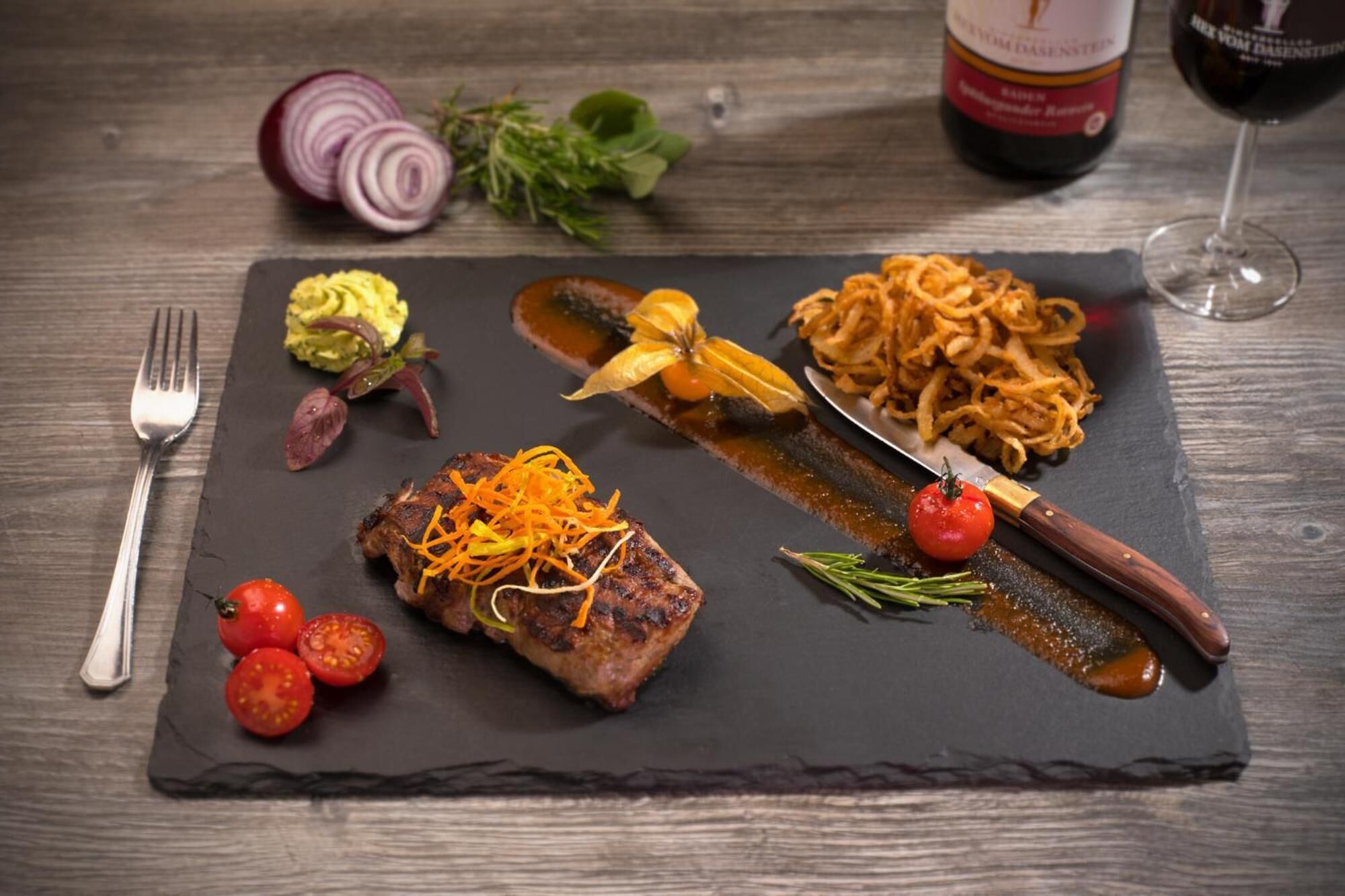 A black slate serving board with a steak topped with carrot shreds, cherry tomatoes, and a knife.