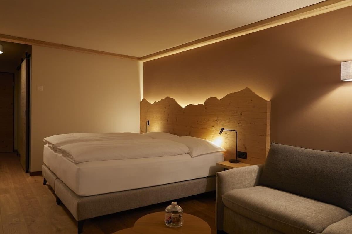 A hotel room with a bed, a nightstand, a lamp, a sofa, and a wooden wall feature.