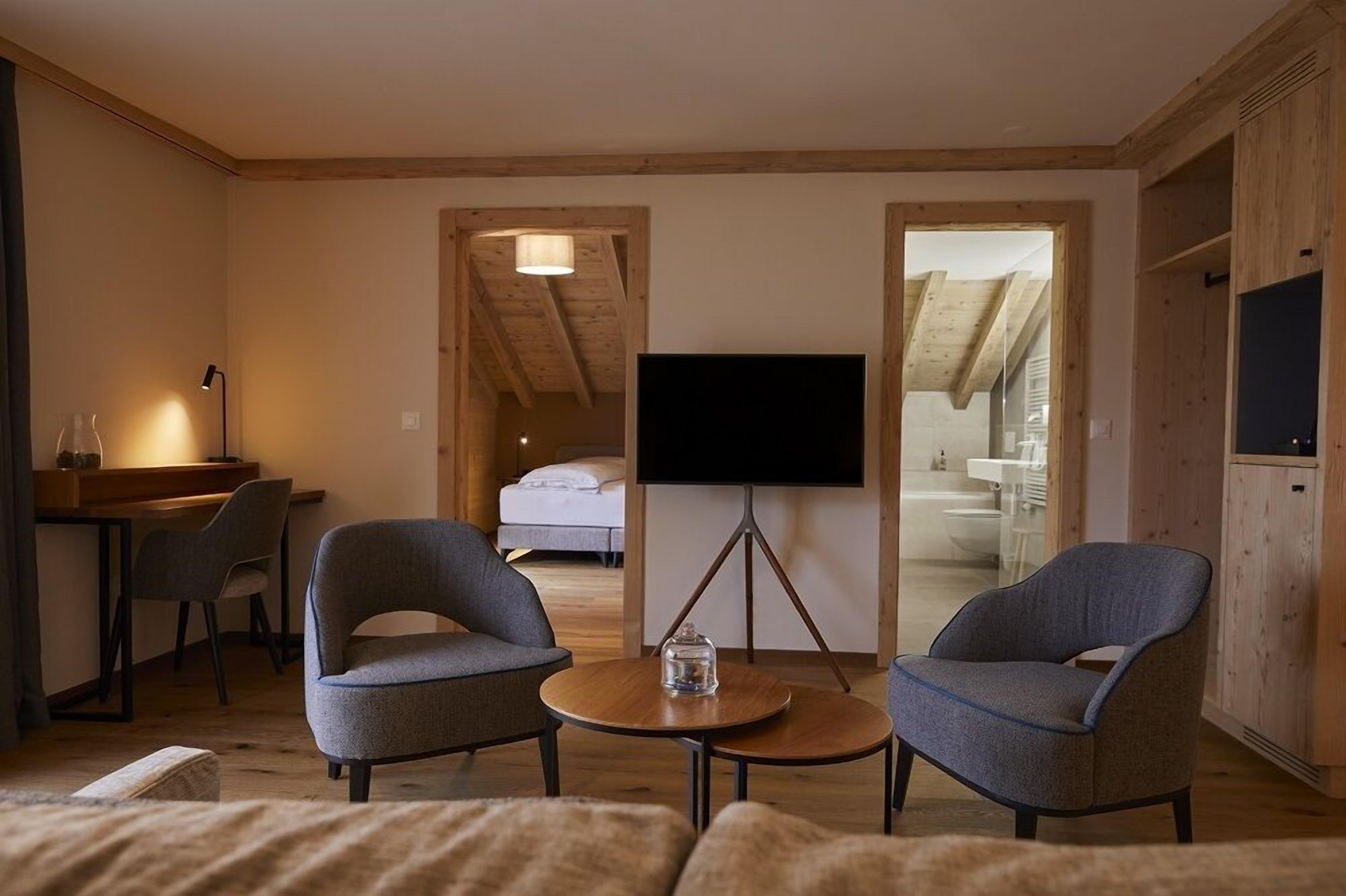 A cozy room with a bed, a TV on a stand, two armchairs, a small table, and a desk with a lamp.