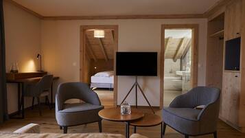 A cozy room with a bed, a TV on a stand, two armchairs, a small table, and a desk with a lamp.