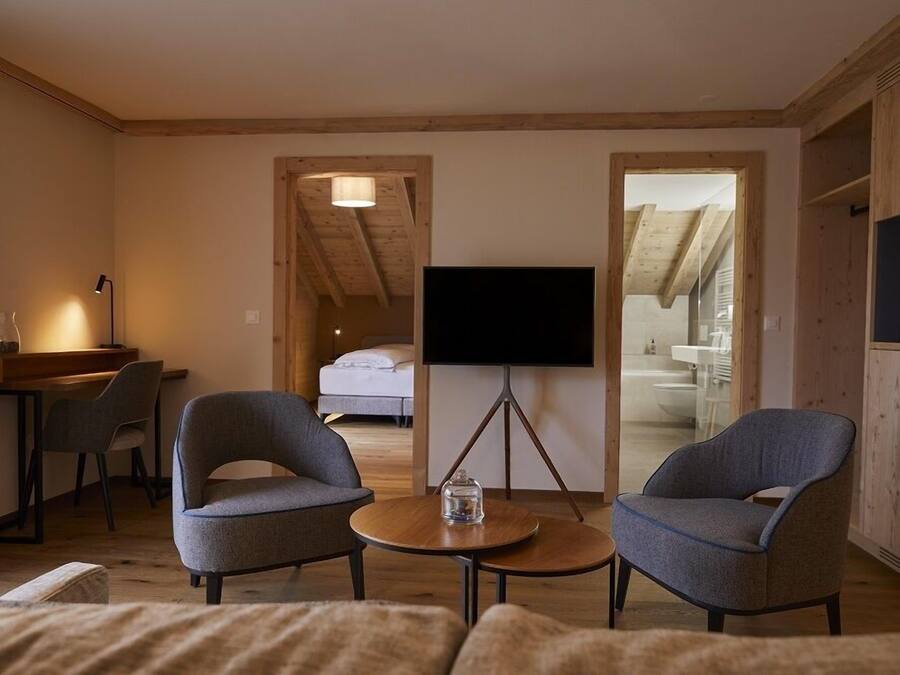 A cozy room with a bed, a TV on a stand, two armchairs, a small table, and a desk with a lamp.