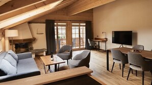 A cozy loft-style room with a wooden ceiling, a fireplace, a sofa, a dining table, and a TV. - Hotel Alpenland (Lauenen)