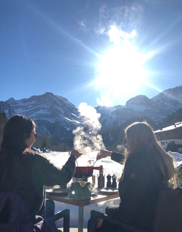 Dos personas sentadas en una mesa con vistas a montañas nevadas y un sol brillante al fondo.