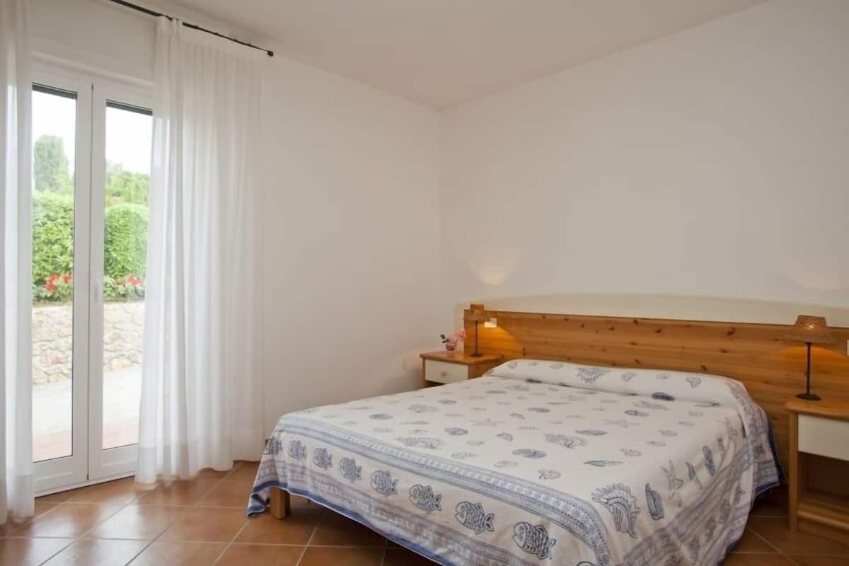 Ein Schlafzimmer mit einem Bett, einem Nachttisch, einer Lampe und einer Schiebetür zu einem Garten.