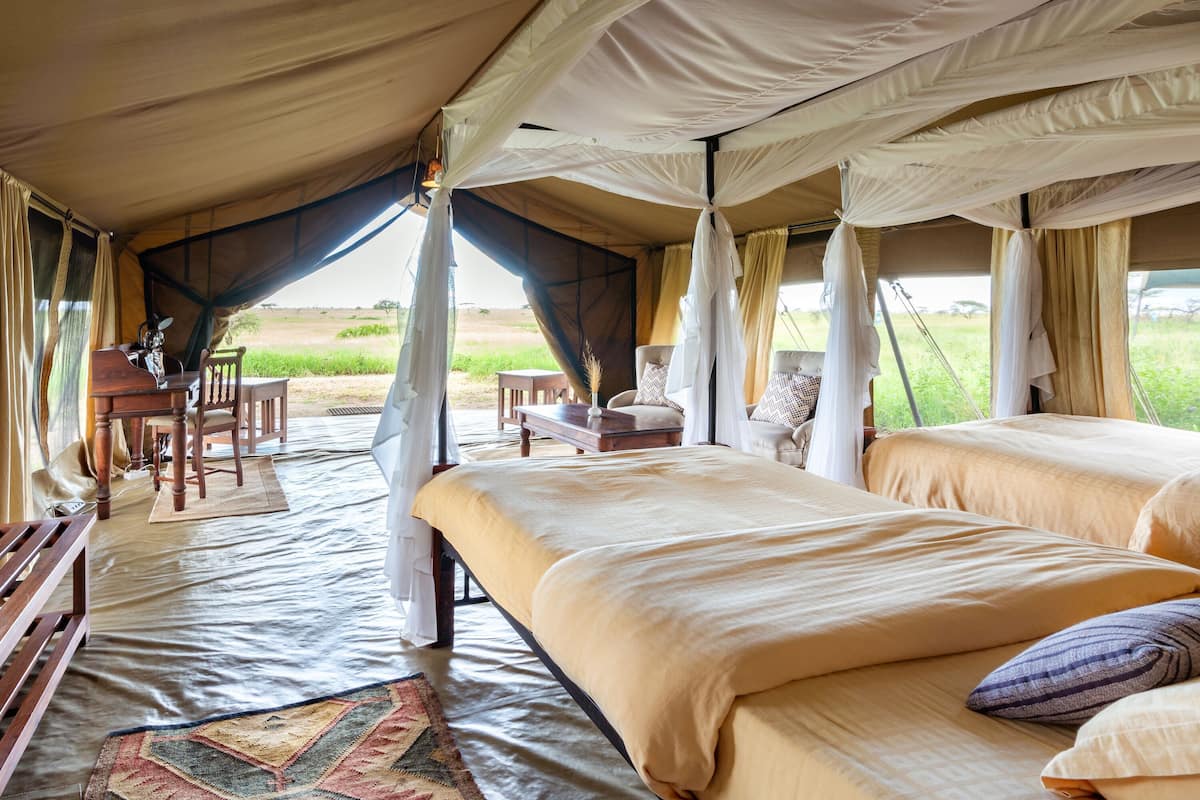 A tented accommodation with two beds, a dining area, and a view of the outdoors.