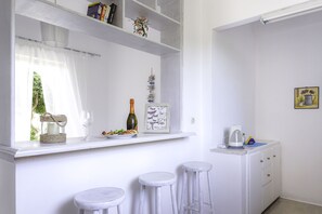 A white kitchen with a window, a shelf with books, a wine bottle, and a framed picture. There are three white stools and a white cabinet with a kettle on top. - Cubaneros Holiday Inn (Zakynthos)