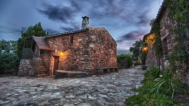 A rustic stone building with a wooden door and a small porch, surrounded by greenery and a cobblestone path.