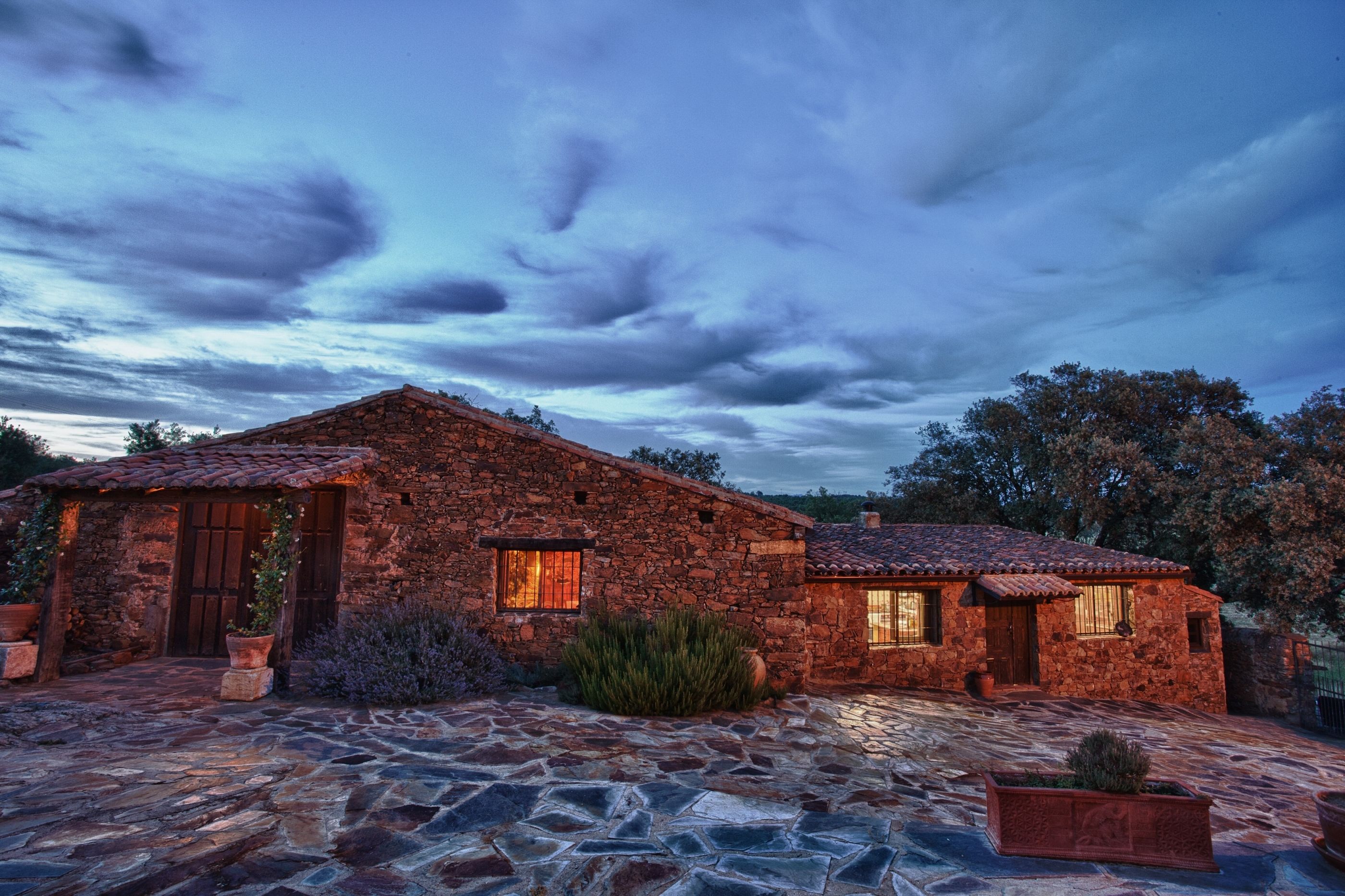 A stone house with a tiled roof, a stone patio, and a large tree in the background.