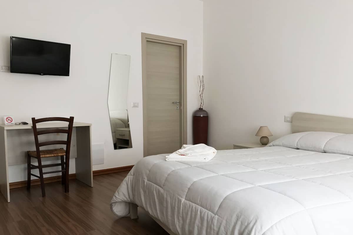 A hotel room with a bed, a desk, a chair, a TV, a mirror, a door, and a nightstand with a lamp.