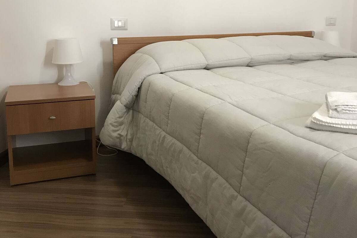 A neatly made bed with a white comforter and pillows, a wooden nightstand with a lamp, and a wall socket.