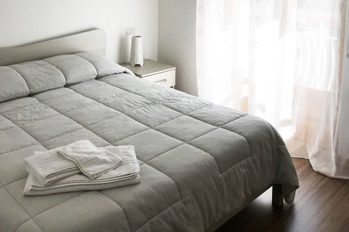A neatly made bed with a grey quilt, a bedside table with a lamp, and sheer curtains with a heart pattern.