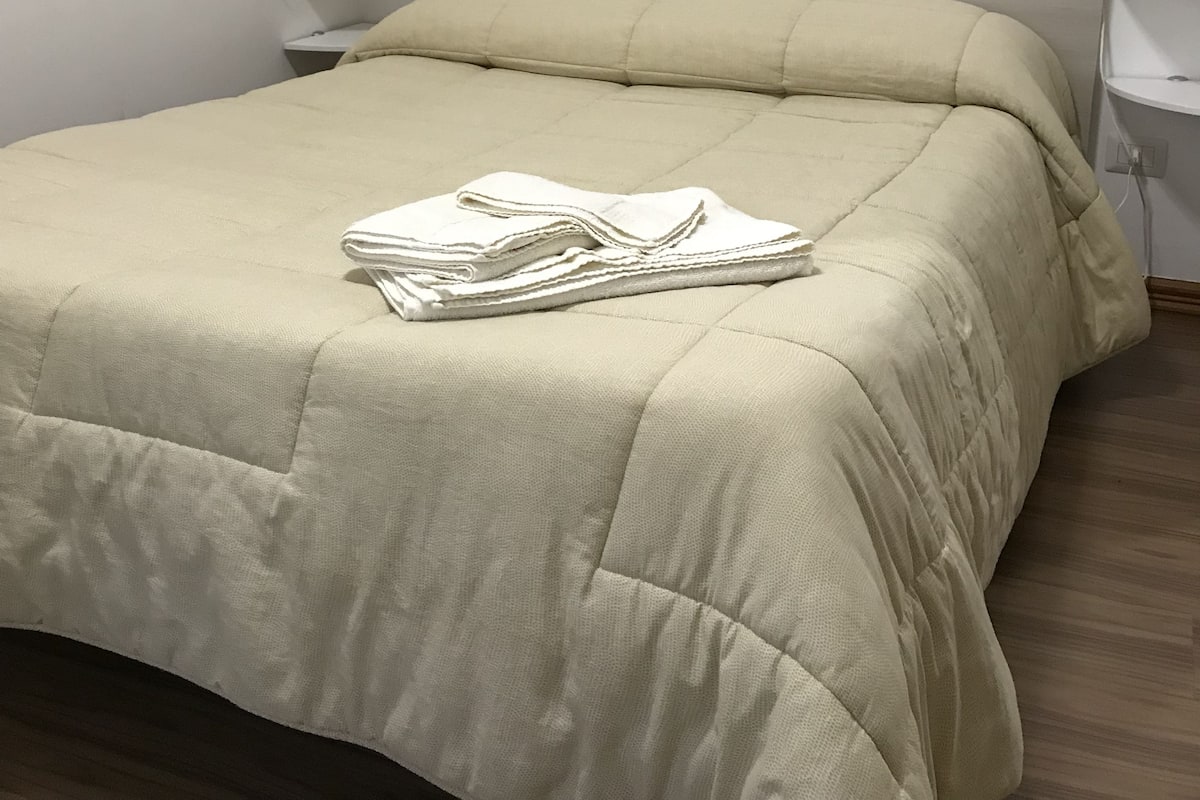 A neatly made bed with a beige comforter and pillows, a bedside table with a lamp, and a wall-mounted power outlet.