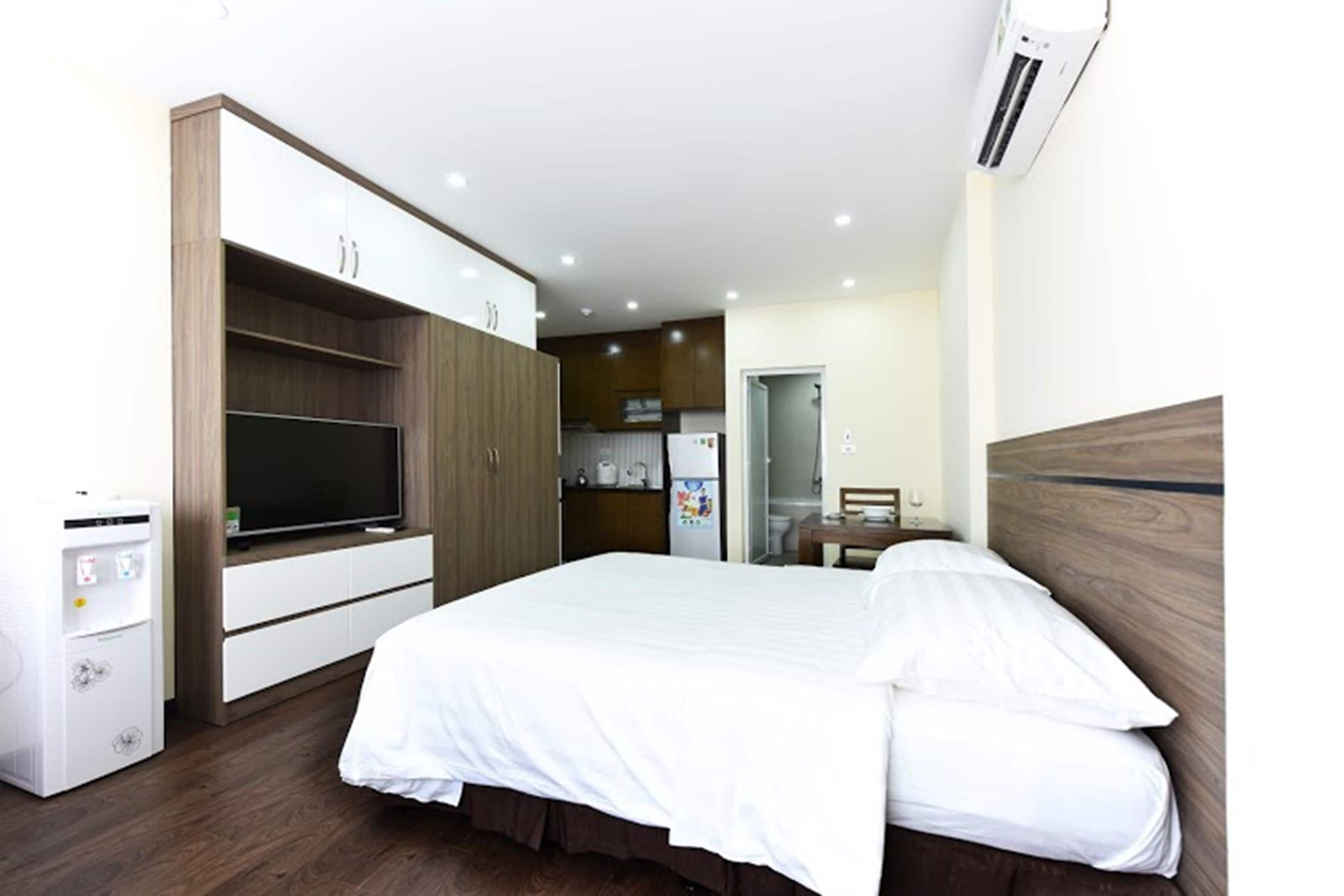 A modern hotel room with a large bed, a TV, a mini-fridge, and a kitchenette in the background.