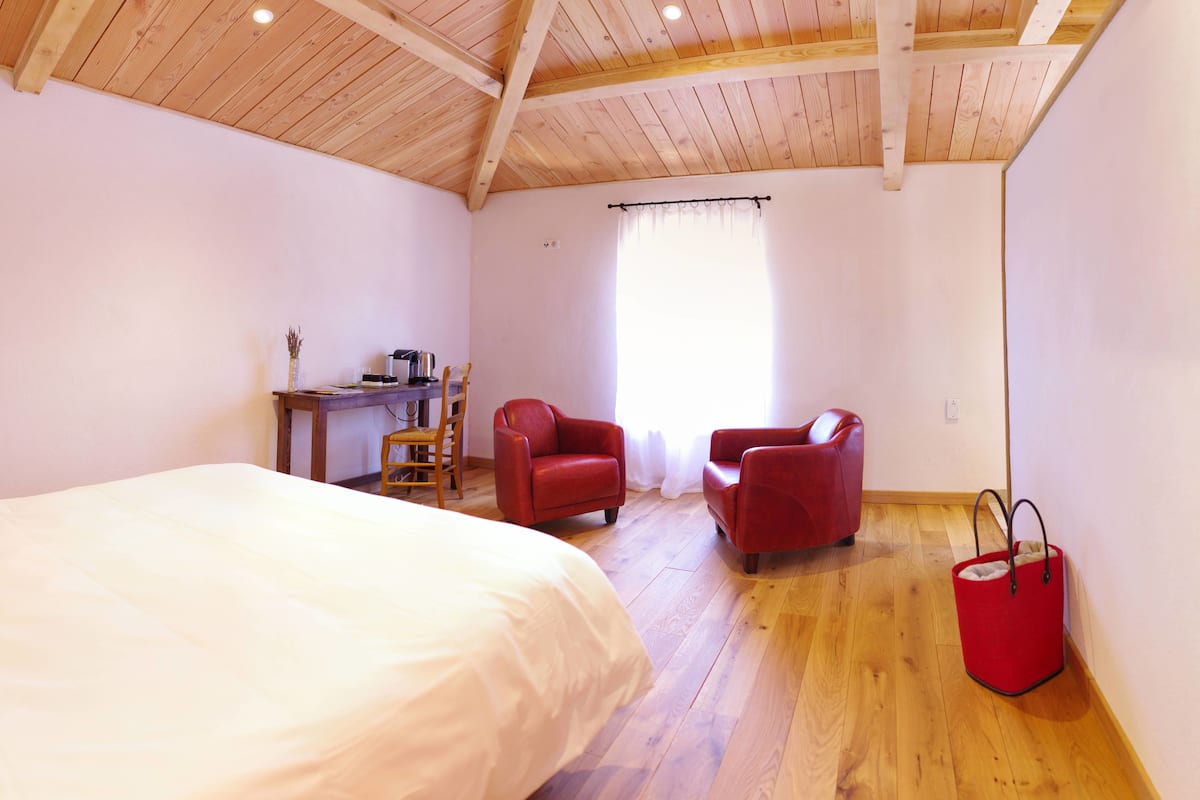 Een slaapkamer met een houten plafond, een bed, een bureau met stoel, en twee rode fauteuils.