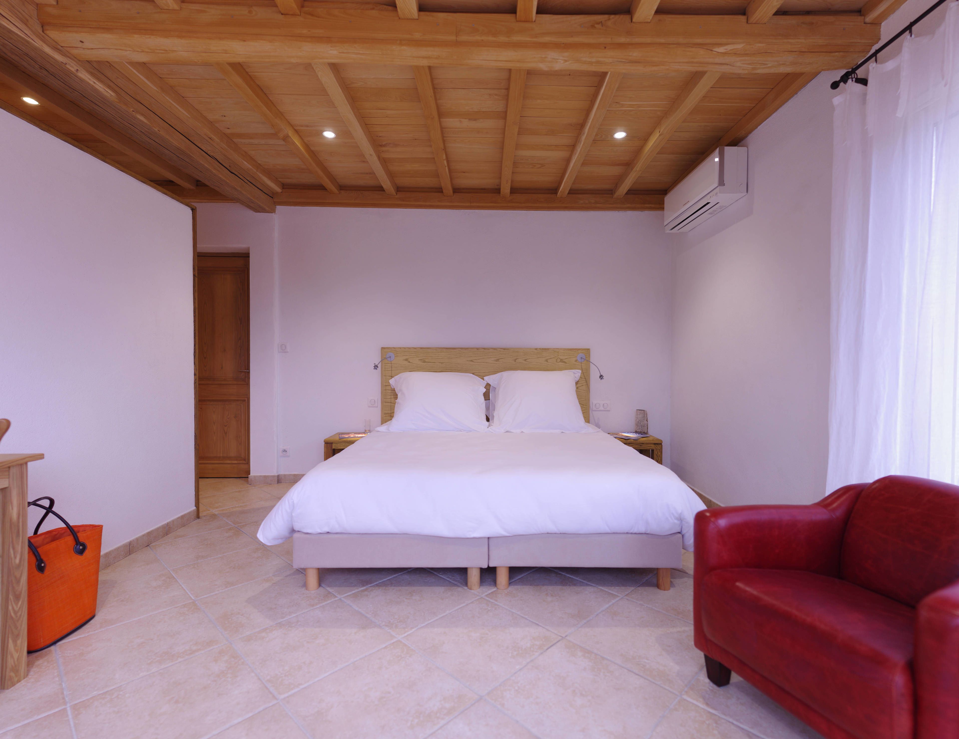 A bedroom with a wooden ceiling, a bed with white bedding, a red chair, and a wooden nightstand.