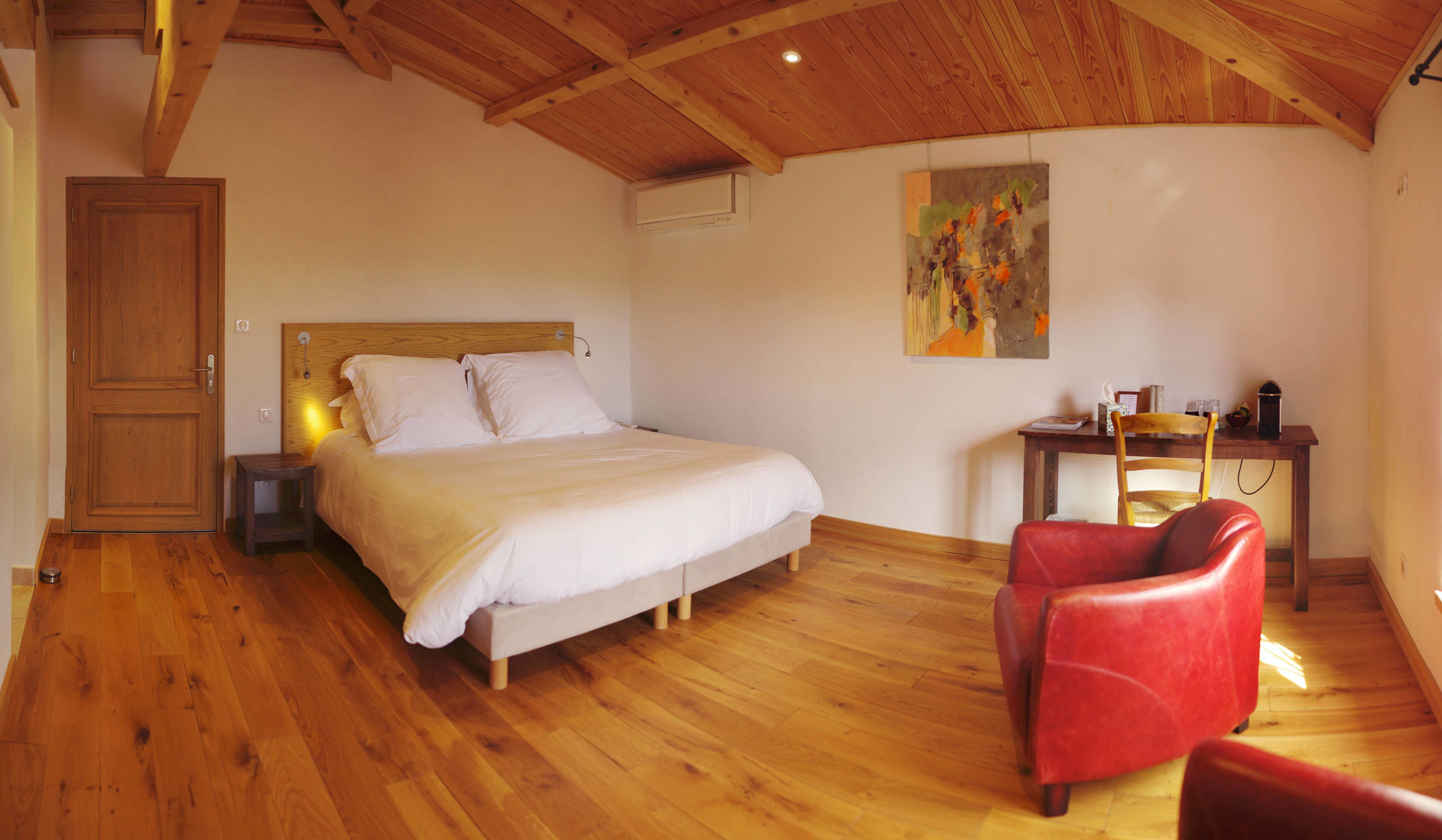 A bedroom with a wooden ceiling, a bed with white bedding, a red chair, and a desk with a chair.