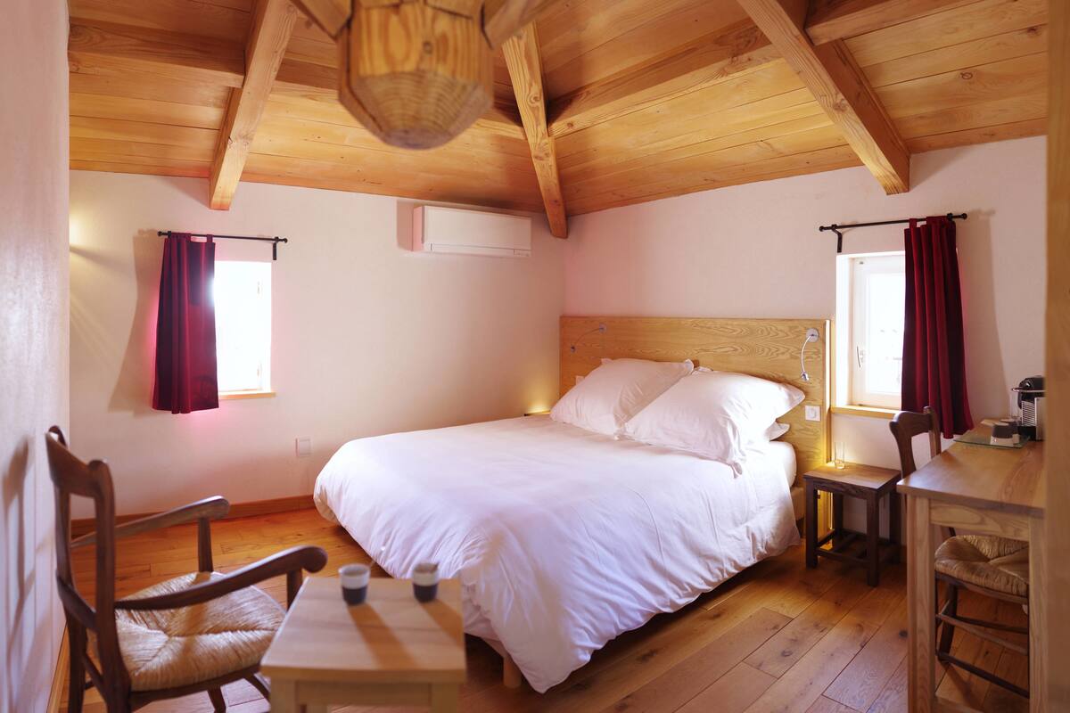 Een gezellige slaapkamer met een houten plafond, een bed met witte lakens, een houten tafel met stoelen, en een klein bureau met een stoel.