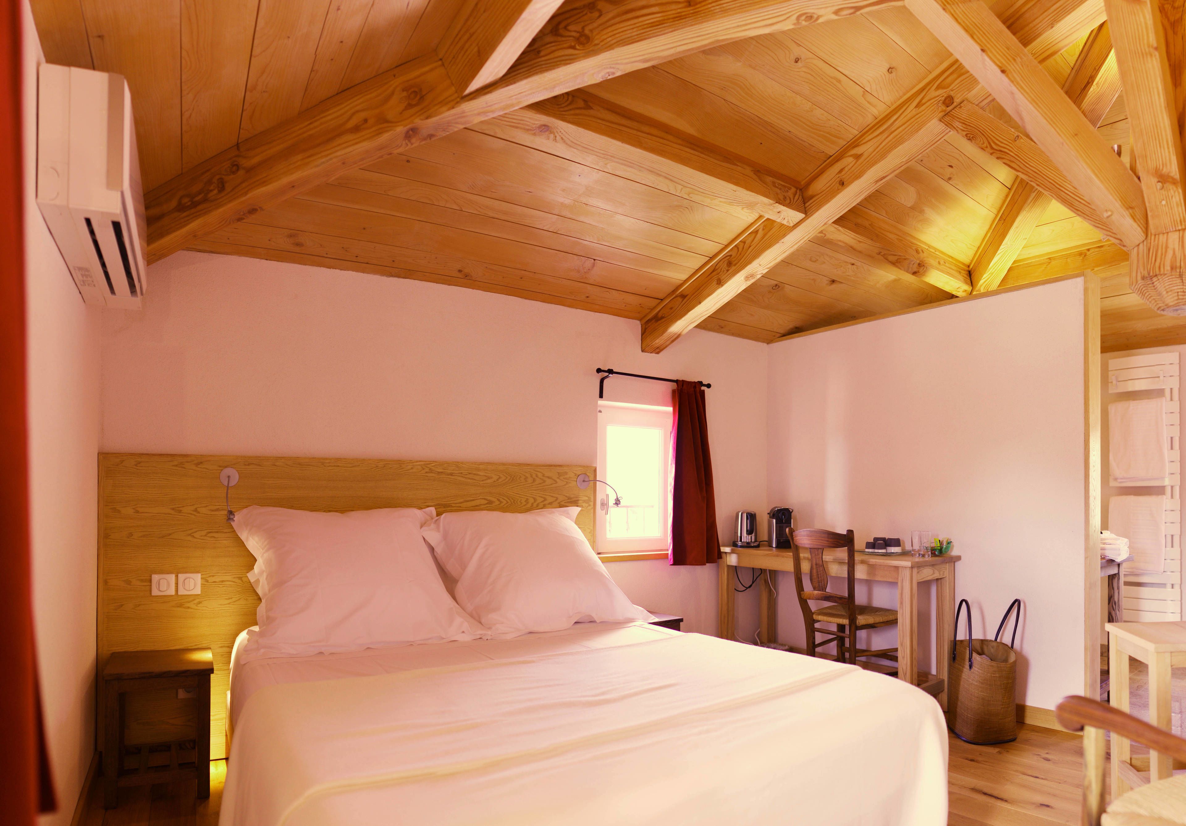 Un dormitorio con techo de madera, una cama con ropa de cama blanca, una mesita con una silla y una ventana con cortinas.