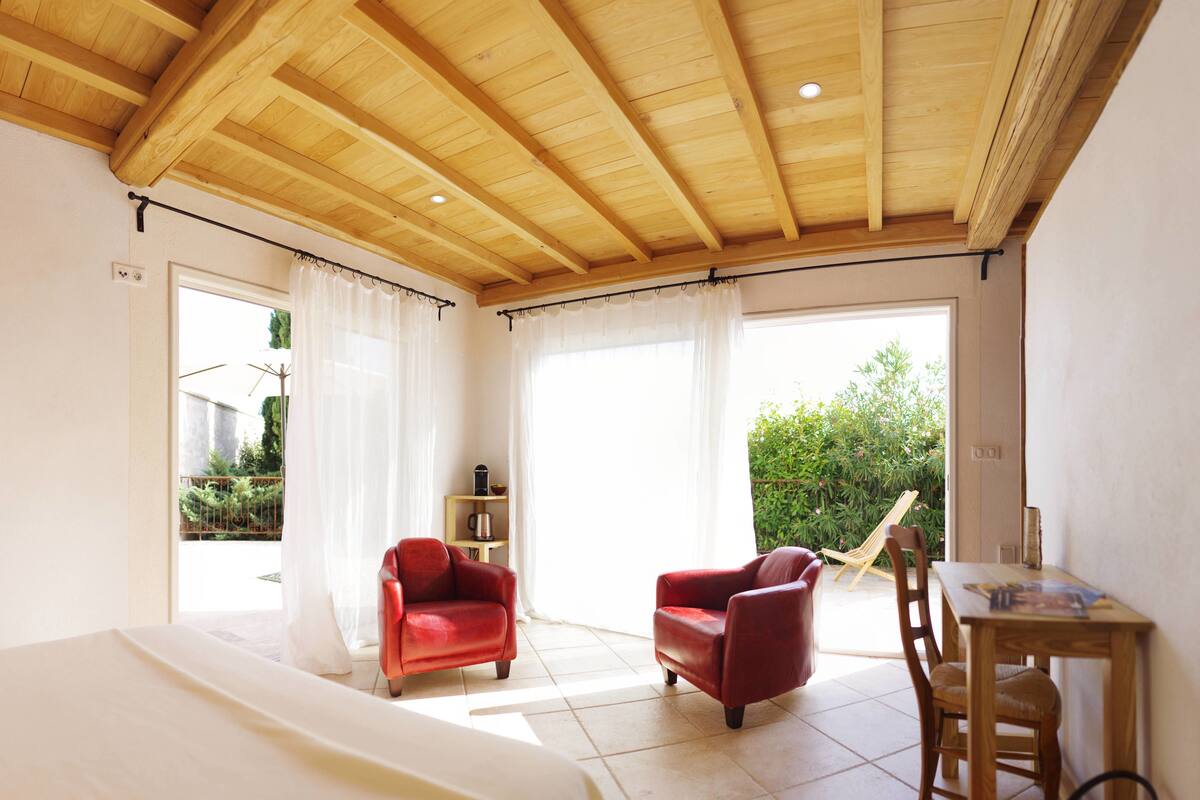 Een slaapkamer met een houten plafond, een bed, een rode stoel, een houten tafel en uitzicht op groen buiten.