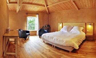 Un dormitorio con techo de madera, una cama con ropa blanca, un escritorio con silla y una ventana con vistas a follaje.