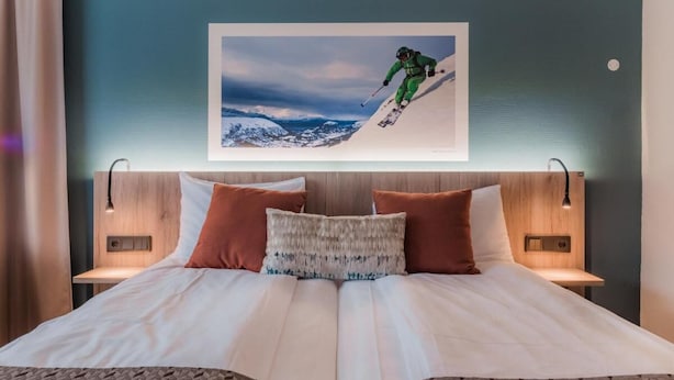 A hotel room with a large bed, two bedside tables, a headboard with a ski-themed artwork, and a nightstand with a lamp. - Nordfjord Hotell - Bryggen (Stad)