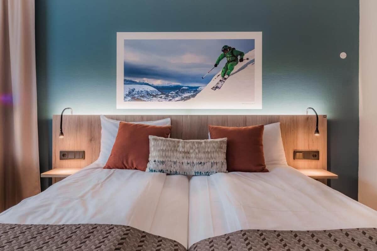 Quarto de hotel com uma cama grande, duas mesas de cabeceira, uma cabeceira com arte decorada com tema de esqui e uma mesinha de cabeceira com uma luminária.