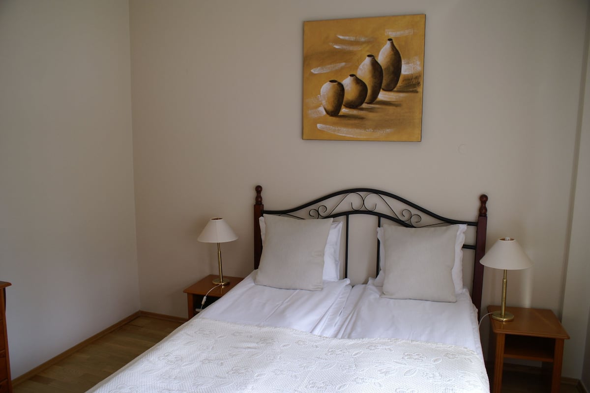 Uma cama bem arrumada com roupa de cama branca, duas mesas de cabeceira com abajures e um quadro na parede.