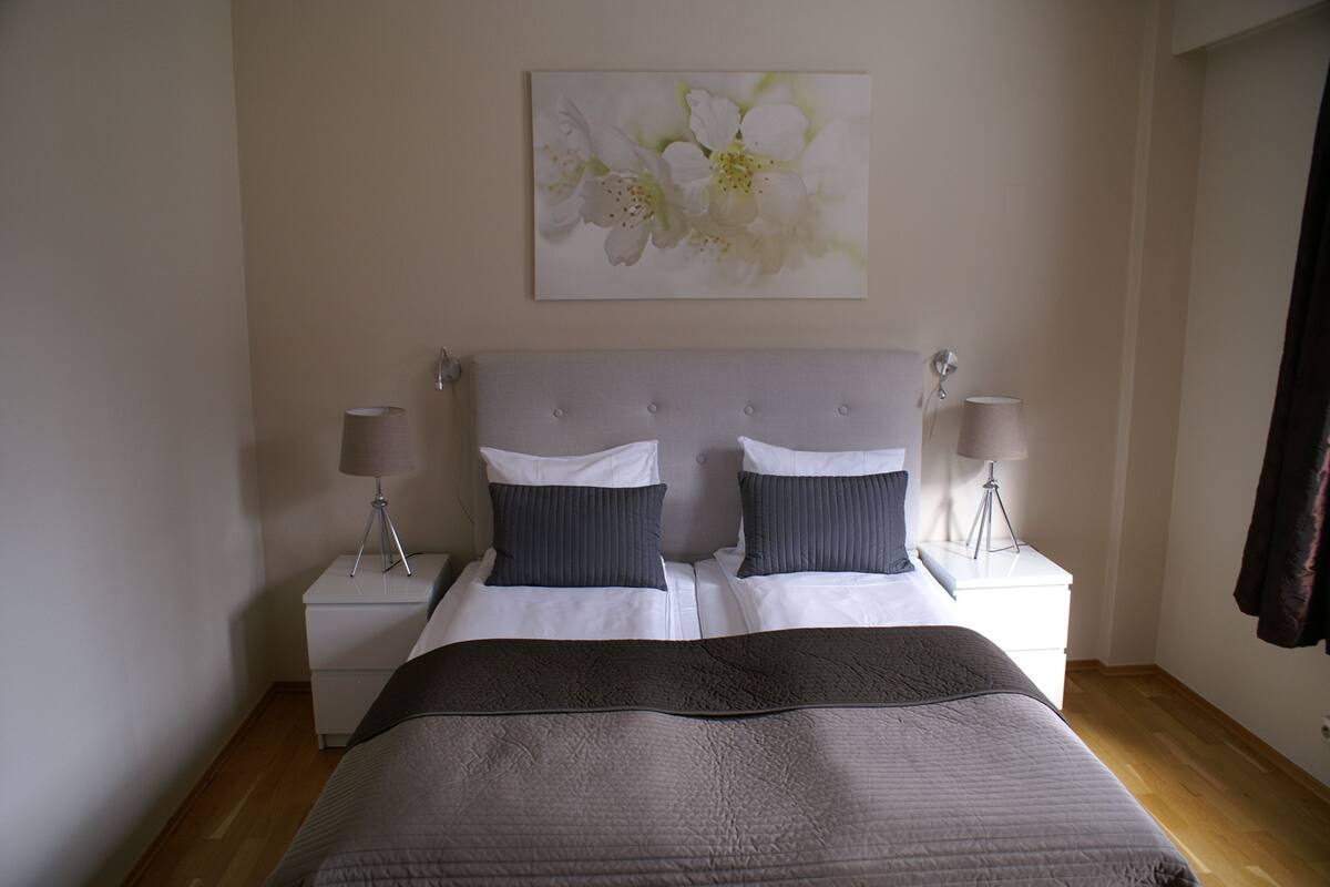 Uma cama de casal com cabeceira cinza, duas mesas de cabeceira com abajures e uma obra de arte floral na parede.