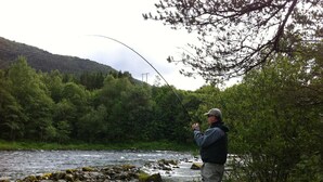 A person fishing by a river with a forested hillside in the background. - Nordfjord Hotell - Bryggen (Stad)