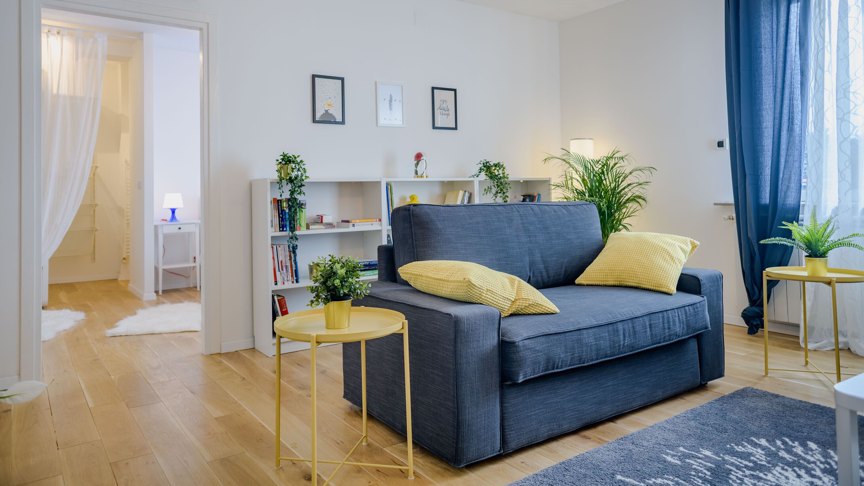 Un salon moderne avec un canapé gris, deux coussins jaunes, une petite table ronde et une bibliothèque remplie de livres et d’objets décoratifs.