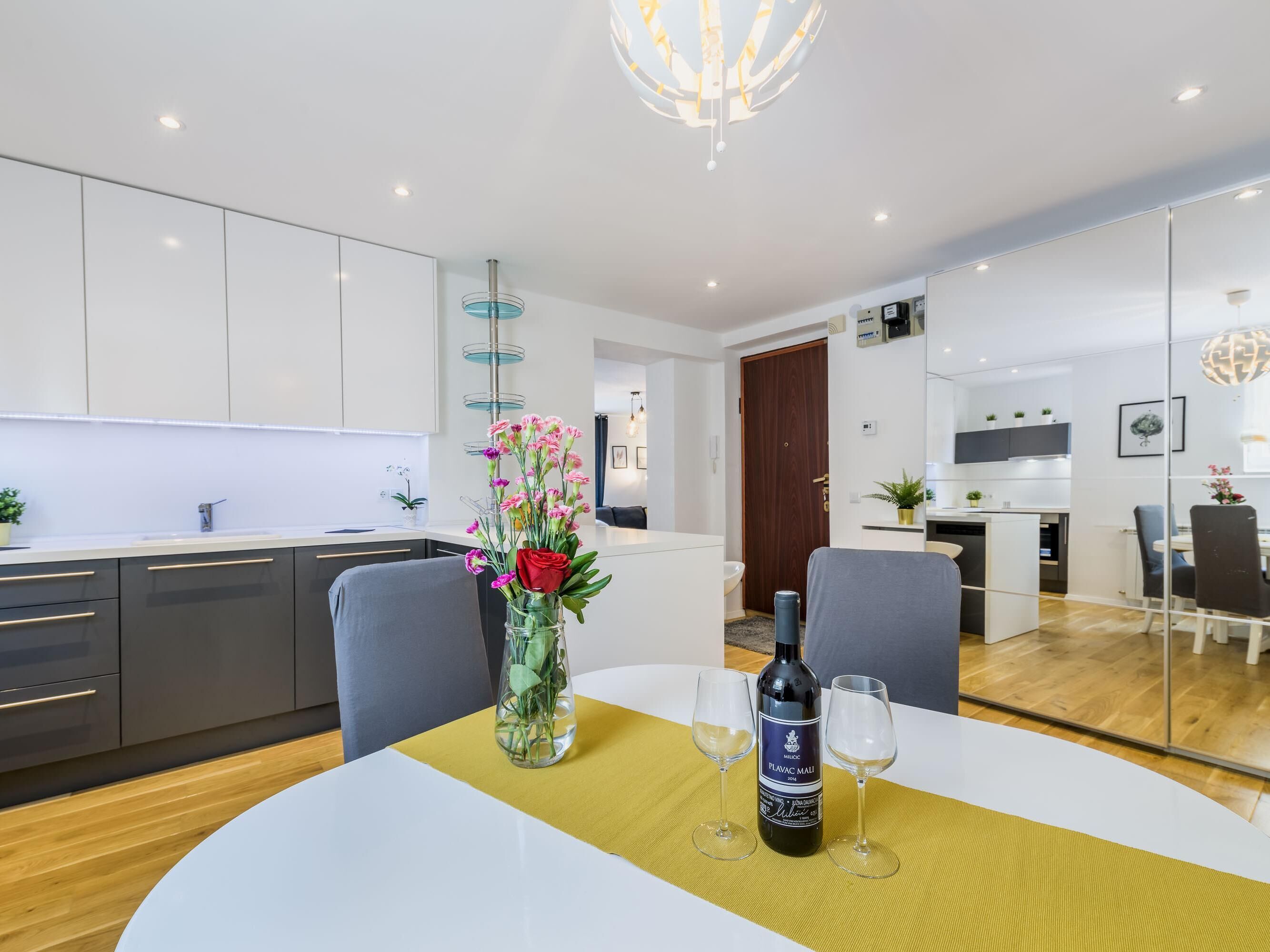 Une cuisine moderne avec des meubles blancs, une table à manger avec deux verres à vin et une bouteille, et un vase rempli de fleurs.