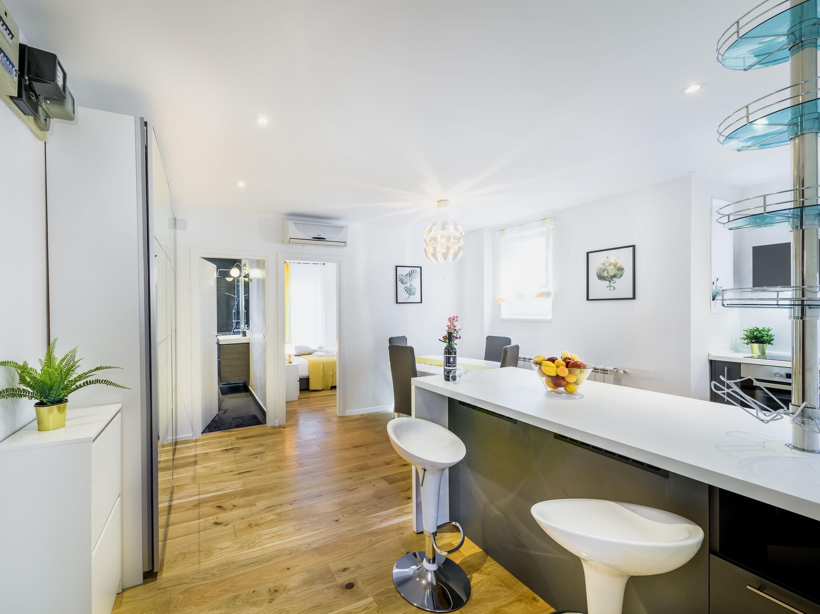 Une cuisine moderne avec un plan de travail blanc, un évier et un coin bar. Le sol est en bois et un couloir mène à une chambre.