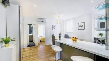 Une cuisine moderne avec un plan de travail blanc, un évier et un coin bar. Le sol est en bois et un couloir mène à une chambre.